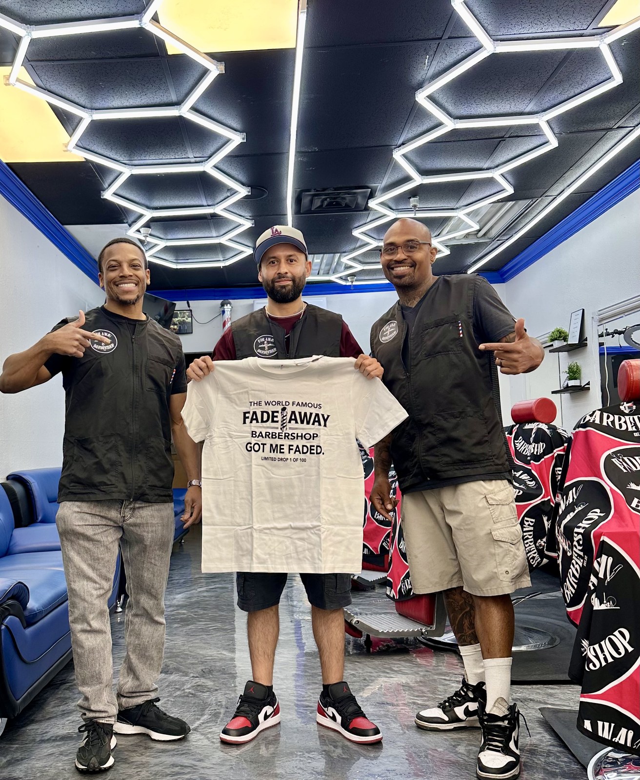 Fade Away Barbershop team holding The World Famous Fade Away Barbershop Got Me Faded limited drop shirt under hexagon LED lights